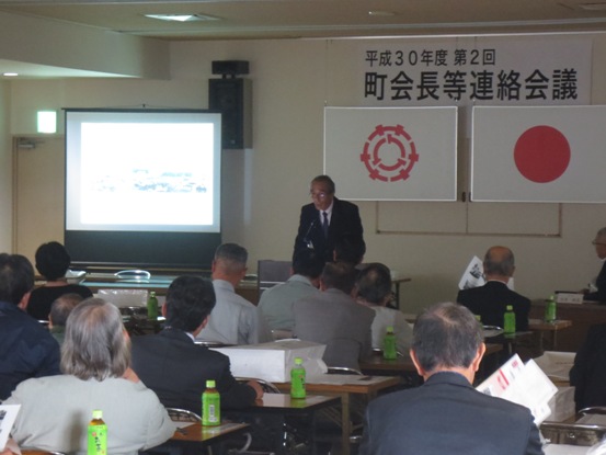 「平成30年度第2回町会長等連絡会議」のボードが吊られている会場で、スクリーンに映し出されたモニターを見つめる関係者たちと、正面に立って発表をしている吉田町会長の写真