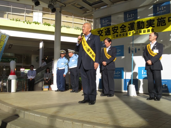 肩に黄色のタスキを下げた市長が、交通安全運動の横断幕が設置された舞台に並び挨拶をしている写真