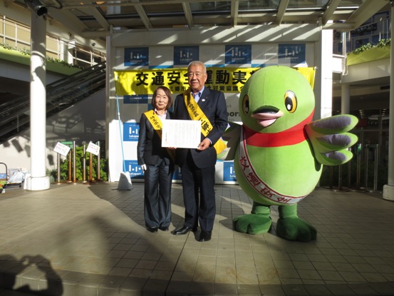 肩に黄色のタスキを下げた市長が、交通安全運動の横断幕が設置された会場で、小林会長やかいちゃんと一緒に並んでメッセージを手に記念撮影をしている写真