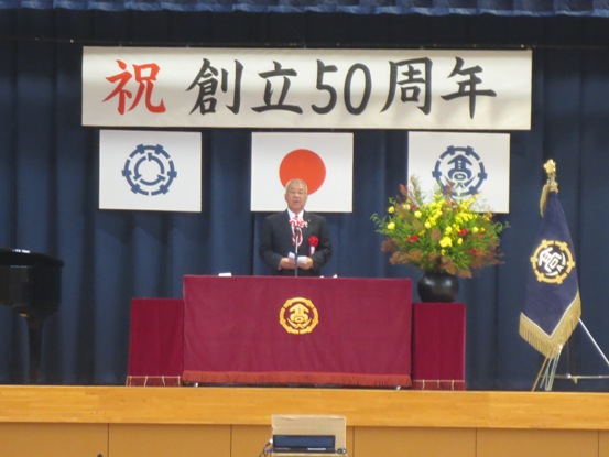 「祝 創立50周年」の吊り看板が設置された舞台上で、演壇に立って話をしている胸章を付けた市長の写真
