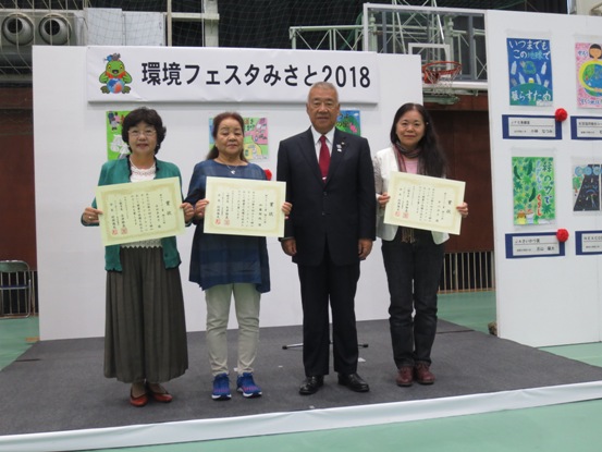 「環境フェスタ2018」のボードの前で、表彰状を持った3名の受賞者と市長が並んで記念撮影をしている写真