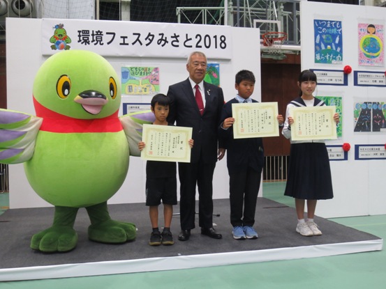 「環境フェスタ2018」のボードの前で、表彰状を持った3名の児童と市長やかいちゃんが並んで記念撮影をしている写真