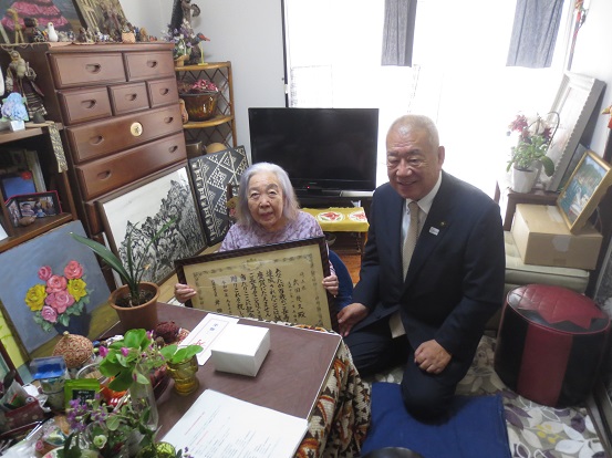 部屋の中で表彰状を持って座る武田幾久さんと、横に座って一緒に記念撮影をしている木津市長の写真