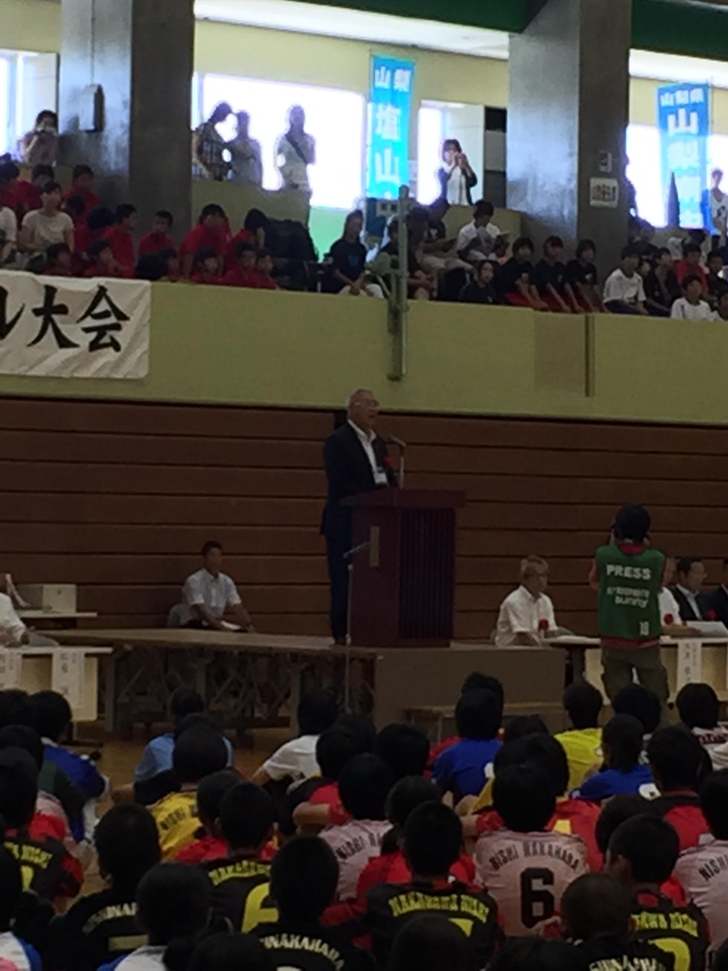 体育館に設置された檀上に立ち挨拶を行っている市長の話を体育座りをして聞いている選手たちの後ろ姿の写真