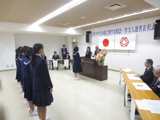 「平成30年度中学生の税に関する標語・作文入選者表彰式」と書かれている横断幕と国旗・三郷市旗が吊り下げられている壁の前で、ピンク・黄色・白の豪華な花々で彩られた花台と並ぶ演台に立ち表彰状の内容を読み上げる三郷市長と、向かい合って立ち話を聞く受賞した女子中学生、後ろに1列に並び表彰の様子を見守る受賞した中学生の写真