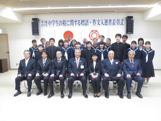 「平成30年度中学生の税に関する標語・作文入選者表彰式」と書かれている横断幕と国旗・三郷市旗が吊り下げられている壁の前で、三郷市長と関係者が受賞した中学生とともに記念撮影をしている写真