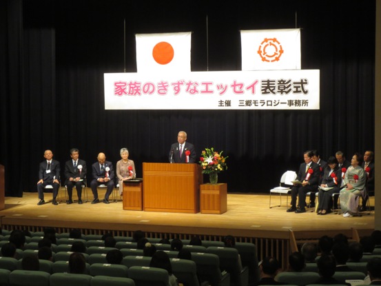 「家族のきずなエッセイ表彰式」と書かれた横断幕と国旗・三郷市旗が吊り下げられているステージ上で、赤・黄色・オレンジ・白の豪華な花々で彩られた花台と並ぶ演台に立ち挨拶の言葉を述べる三郷市長と、ステージ上に置かれたパイプ椅子に座り話を聞く胸章を付けた来賓者の写真