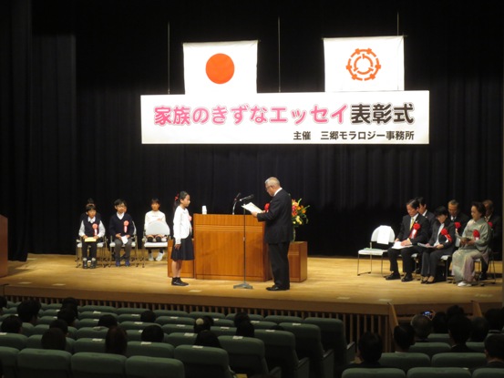 「家族のきずなエッセイ表彰式」と書かれた横断幕と国旗・三郷市旗が吊り下げられているステージ上で、演台の前に立ち表彰状の内容を読み上げる三郷市長と向かい合って立ち話を聞く受賞者の女の子の写真