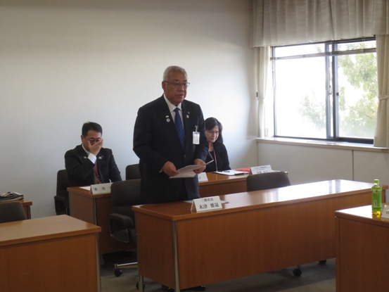 席札が置かれている席を立ち資料を手に持ちながら挨拶の言葉を述べる三郷市長と、後ろの席に座って話を聞く関係者の写真
