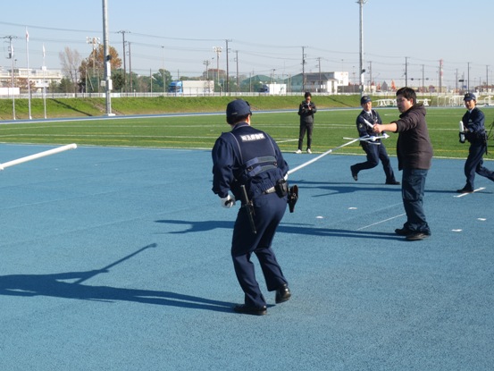 男性が警官に向かって左手で持ったナイフを向けており、3名の警察官が白い棒を持って男性に立ち向かっている緊迫したテロ対処訓練の様子