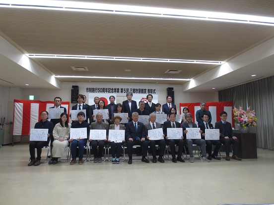 紅白の横断幕の前に多くの受賞者が3列に並んで表彰状を持っているなか、前列の市長を中心に記念撮影をしている写真