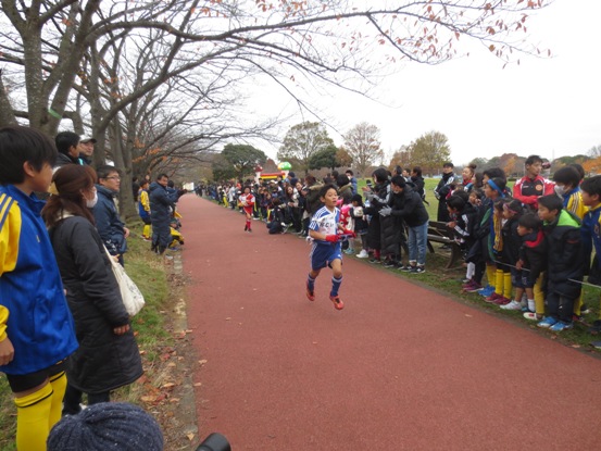 赤く塗装された公園の遊歩道を走る子どもと、遊歩道の両脇から声援を送る関係者たちの写真