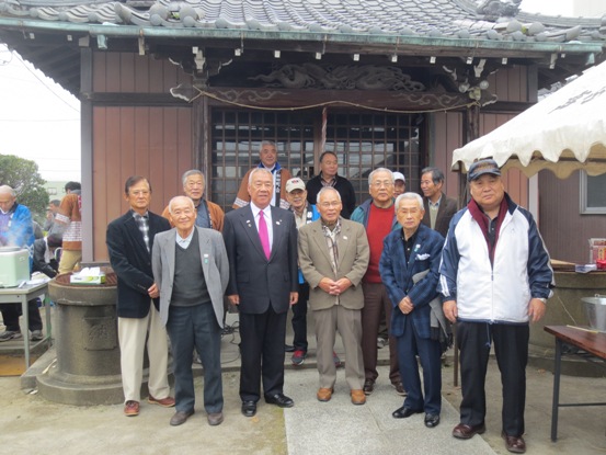 瓦屋根の建物の周りで法被を着て作業をする関係者と、市長と一緒に並んで記念撮影をしている関係者たちの写真