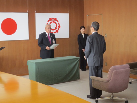 日本国旗と三郷市旗が設置された部屋で、緑の机を挟んで市長と関係者が向かい合っている写真