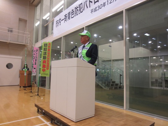 「防犯活動実施中」ののぼりが立てられたガラス張りの室内に演台が設置され、緑の帽子とベストを着用した市長が演台に立って挨拶をしている写真