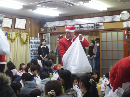 たくさんの子どもたちが座っている部屋で、サンタの衣装を着た市長が、白い大きな袋に手を入れながら子どもたちに微笑んでいる写真