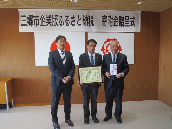 「三郷市企業版ふるさと納税寄附金贈呈式」と書かれた横断幕と国旗、三郷市章が壁に掛けられた部屋で、（左から）人材開発部取締役部長綿貫亮様、代表取締役島田秀貴様、三郷市長が並び記念撮影をしている写真