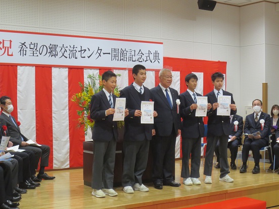 「祝 希望の郷交流センター開館記念式典」と書かれたプレートと紅白の幕が設置された会場で、命名した瑞穂中学校の4人の生徒と三郷市長が並び記念撮影をしている写真