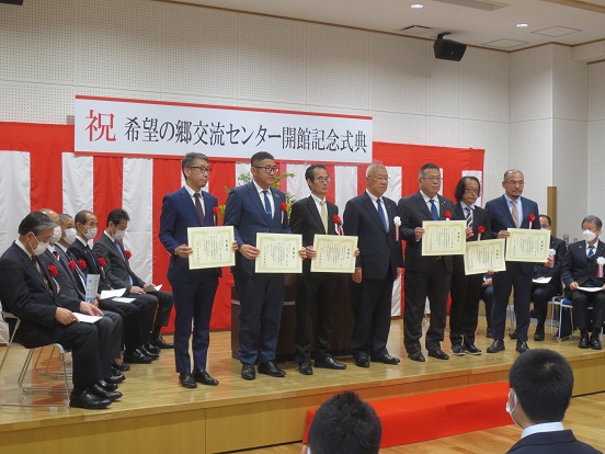 「祝 希望の郷交流センター開館記念式典」と書かれたプレートと紅白の幕が設置された会場で、6人の事業者の方々と三郷市長が並び記念撮影をしている写真