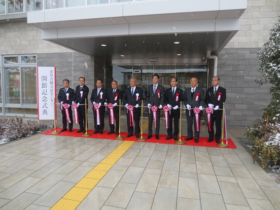 「希望の郷交流センター開館記念式典」と書かれた看板が立っている入口前で三郷市長と関係者の方々がレッドカーペットに並び、テープカットをしている写真
