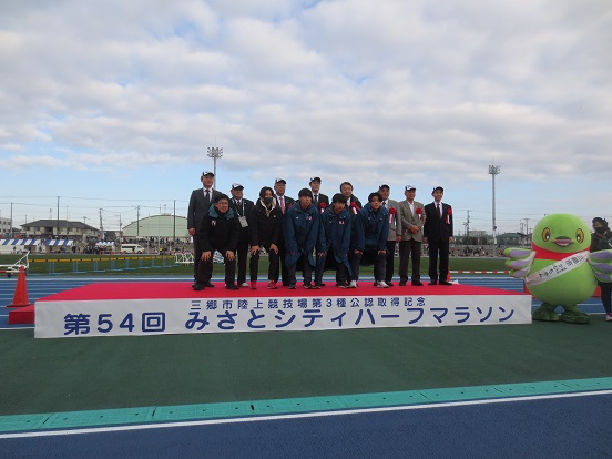「第54回みさとシティハーフマラソン」と書かれた台の上に三郷市長と関係者の方々、3人の日本大学陸上競技部の選手が乗り、台から降りているかいちゃんも一緒に記念撮影をしている写真