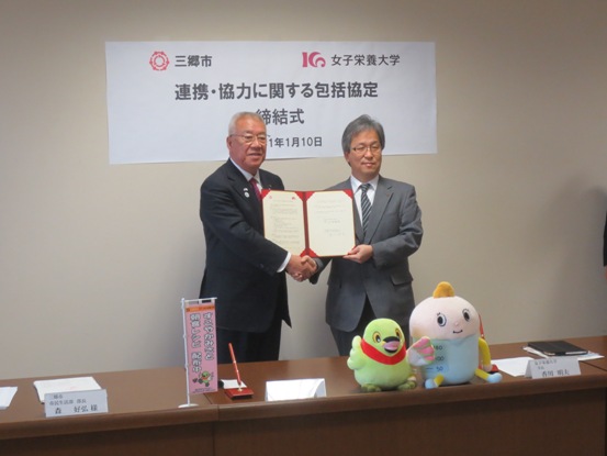 「連携・協力に関する包括協定」と書かれた紙が壁に貼られている部屋で、関係者と一緒に協定書を持って握手を交わしている市長の写真
