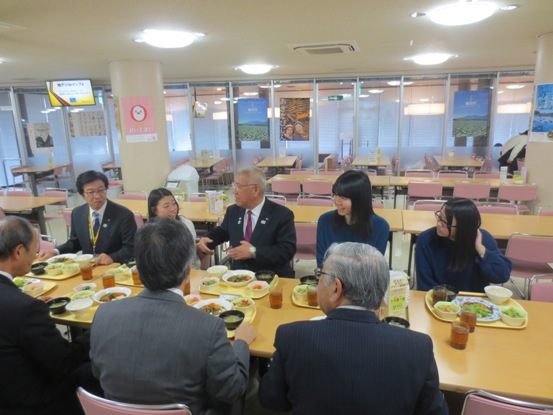 長テーブルにピンク色の椅子が等間隔に設置されている食堂で、市長や関係者、学生のかたと一緒に座って学食を前に会話をしている様子の写真
