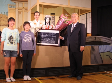 演者が花束と額入りの写真を市長と一緒に持ち児童と一緒に並んでいる記念写真