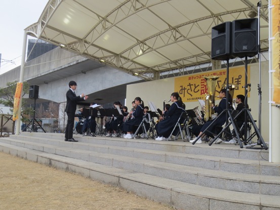 屋外のステージ上で男性指揮者と吹奏楽部の生徒たちが演奏を披露している写真