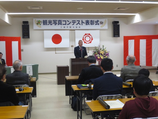 「観光写真コンテスト表彰式」の吊り看板や紅白の横断幕が設置された部屋で、等間隔に並べられた長机に2名ずつの受賞者が座るなか、正面にたつ市長が挨拶をしている写真