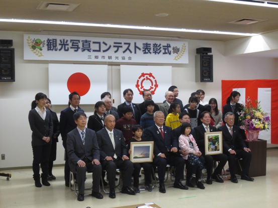 「観光写真コンテスト表彰式」の吊り看板の下で、様々な年代の受賞者と一緒に並んで記念撮影をしている市長の写真