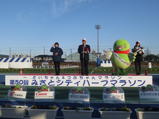 「第50回みさとシティハーフマラソン」のパネルが設置された会場で、かいちゃんや関係者が横に立っているなか、ステージ上で挨拶をしている市長の写真