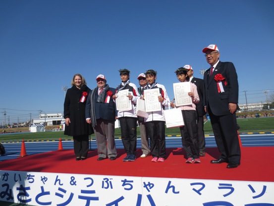 レッドカーペットの舞台がセットされた陸上競技場で、駐日ギリシャ大使や市長が小中学生の入賞者と一緒に舞台に立って記念撮影をしている写真