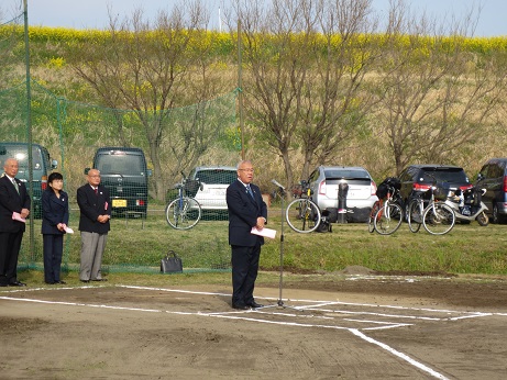 江戸川第3運動公園のグラウンドに立ち設置されたマイクに向かって開会式のあいさつの言葉を述べている三郷市長と、後ろの緑のネット付近に立って話を聞く関係者3名の写真