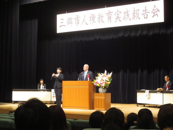 「三郷市人権教育実践報告会」の吊り看板が設置された舞台上で、手話通訳の女性が横に立って手を動かしているなか、演壇に立って挨拶をしている市長の写真