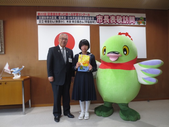 壁に「第22回図書館を使った調べる学習コンクール市長表敬訪問」の紙が貼られている部屋で、制服姿で学用品を手に持っている女子生徒を中心に、市長とかいちゃんが並んで記念撮影をしている写真