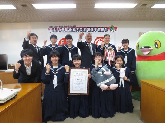 壁に「 平成30年度全国大会出場選手激励会」の紙が貼られている部屋で、制服姿の女子生徒たちが横2列に並んでおり、1列目の女子生徒は表彰状やつぶちゃんマスコットを手に持ち、2列目に立っている市長や関係者、かいちゃんと一緒に記念撮影をしている写真