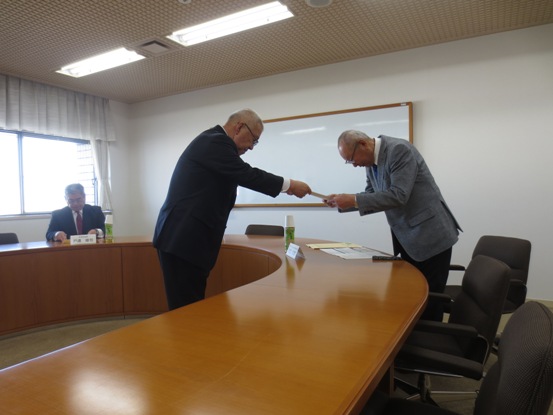 Uの字型のテーブルが設置されている部屋で、市長がテーブル越しに立っている男性に委嘱書を渡している写真