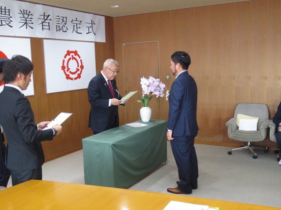 壁に認定式の紙が貼られている部屋で、緑色のクロスが敷かれたテーブル越しに市長から藍色のスーツ姿の男性へ認定書が交付される様子の写真