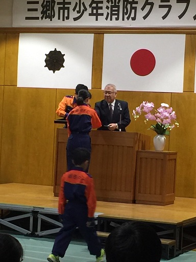 「三郷市少年消防クラブ入団式」と書かれた横幕と国旗が飾られた室内で、演壇を挟んでオレンジに紺色の作業着を着用した団員と向かい合って立っている市長が少年消防手帳を手渡している様子の写真
