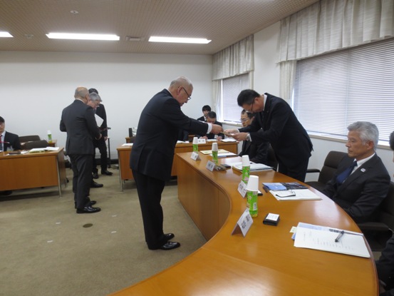 カーブのかかった長テーブルが設置されている部屋で、スーツ姿の関係者たちが着席しているなか、一名の男性へテーブル越しに委嘱書を渡している市長の写真