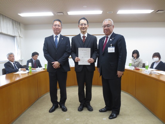 諮問書を持っている男性を真ん中に、市長を含め3名で並んで記念撮影をしている写真