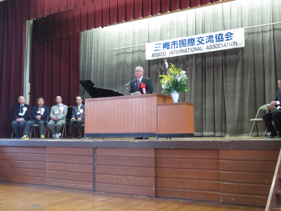 「三郷市国際交流協会」の吊り看板が設置された舞台上で、胸章をつけた市長が演壇に立って挨拶をしている写真