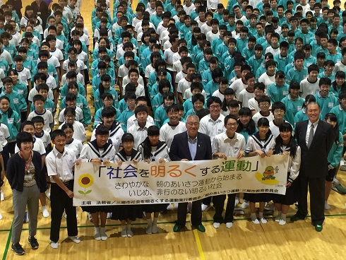 体育館内で「社会を明るくする運動」と書かれた横断幕を市長を中央に7名の生徒が持ち、両端に2名の先生、後ろに体操着を着た生徒たちが整列して並んでいる写真