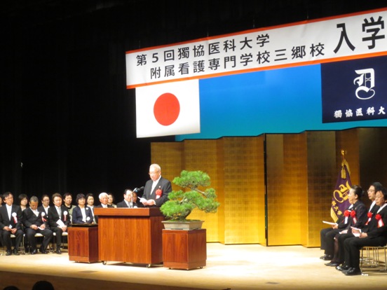 上部には「第5回獨協医科大学附属看護専門学校三郷校入学式」の吊り看板、花台には盆栽、演台の両側には多くの関係者たちが着席している舞台上で、演壇に立っている市長が祝辞を述べている様子の写真