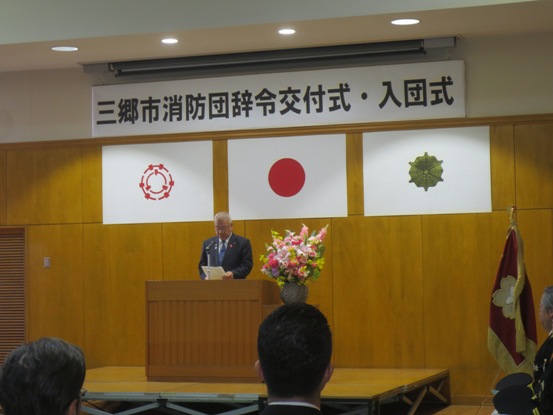 壁に「三郷市消防団辞令交付式・入団式」と書かれたボードが設置されている部屋で、演壇に立つ市長が資料を手に挨拶をしている写真
