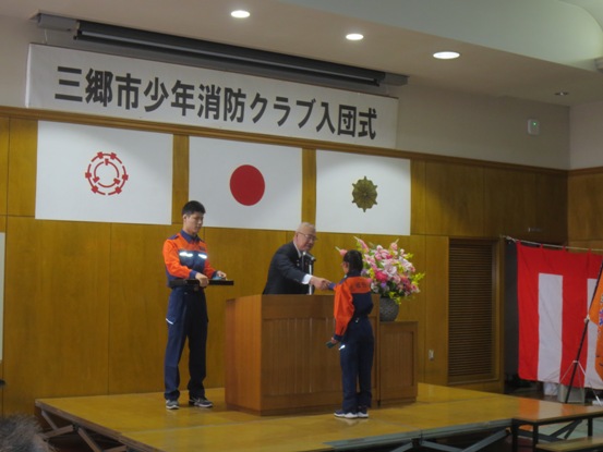 壁に「三郷市消防団辞令交付式・入団式」と書かれたボードが設置されている部屋で、演壇に立つ市長と向かいに立つ児童が握手を交わしている写真