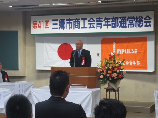 「第41回三郷市商工会青年部通常総会」の吊り看板が設置された部屋で、スーツ姿の関係者たちが着席しているなか、演壇に立つ市長が挨拶をしている写真