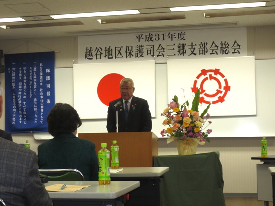 「平成31年度越谷地区保護司会三郷支部会総会」の紙や花台が設置されている部屋で、演壇に立つ市長が挨拶をしている写真