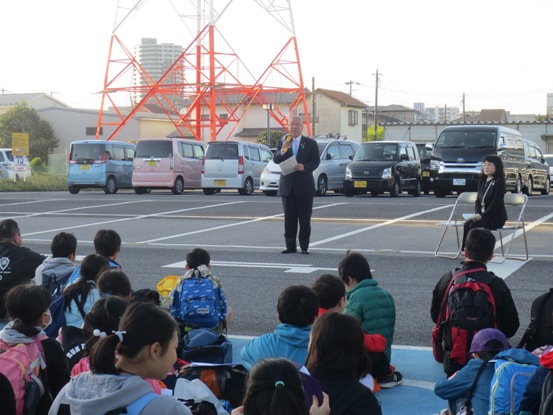 駐車場内の一角で、多くの子ども達が座っている中、正面に立つ市長がマイクを手に挨拶をしている写真